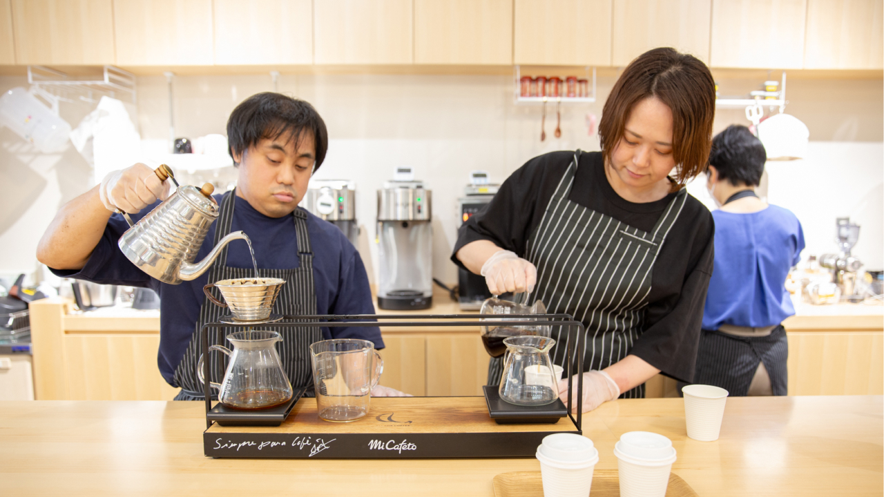 男性1人、女性1人が木目調のカフェでコーヒーを注いでいる様子の写真